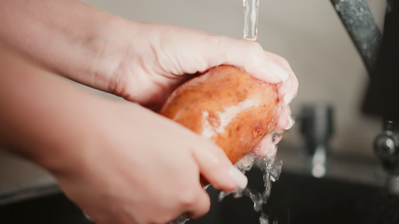 Close up hands of fair skinned person washing fresh brown potato under running water from faucet, emphasizing food hygiene, freshness, and kitchen preparation in clean soft blur environment