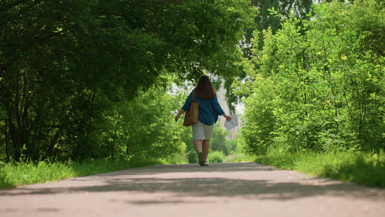 Back view of happy lady walking along lush green alley under sunlight, dancing playfully while holding embroidered fabric in hand, with fabric bag, residential buildings faintly visible ahead