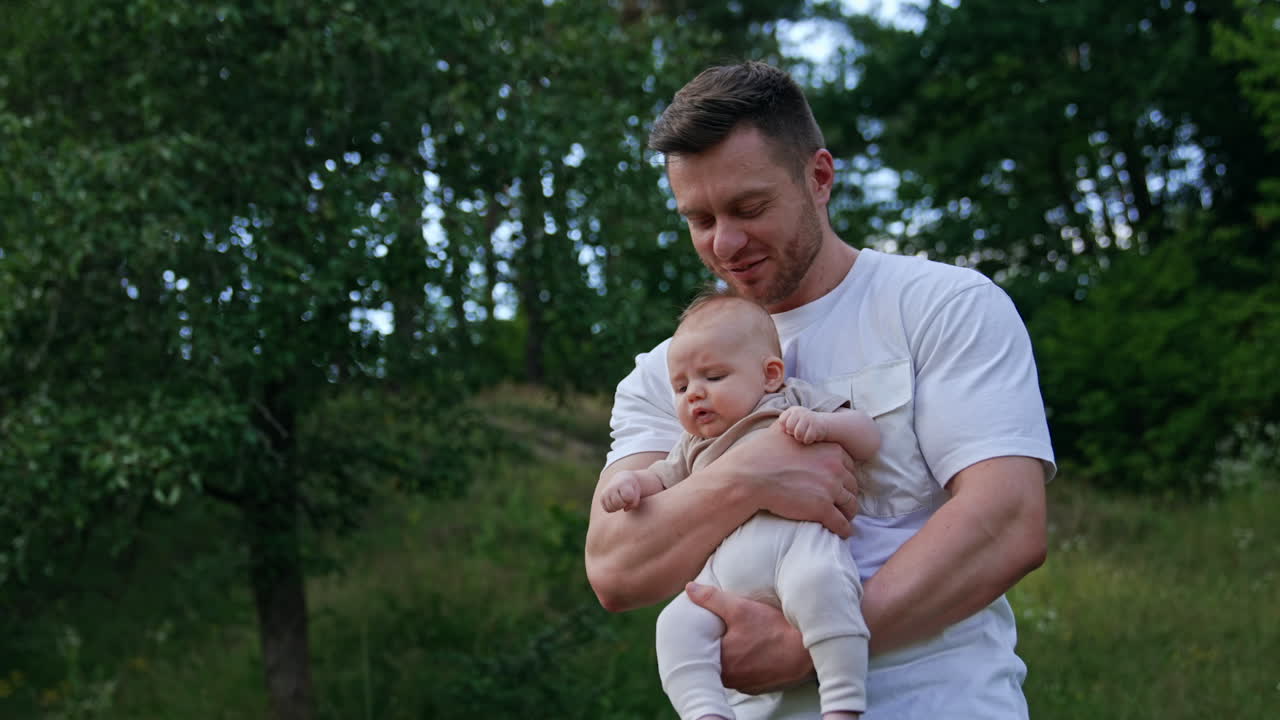 Man waving his baby boy in hands. Caring happy dad kisses child's head. Blurred nature at backdrop.