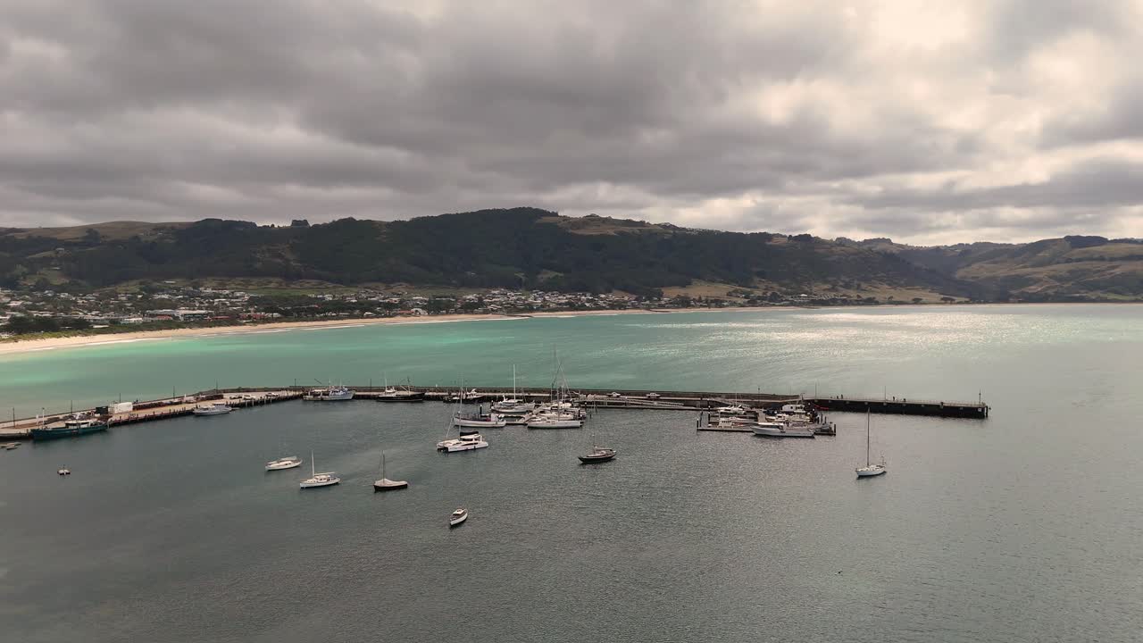 Drone footage captures Apollo Bay harbor with boats under cloudy skies. Calm waters and distant hills create a serene coastal scene