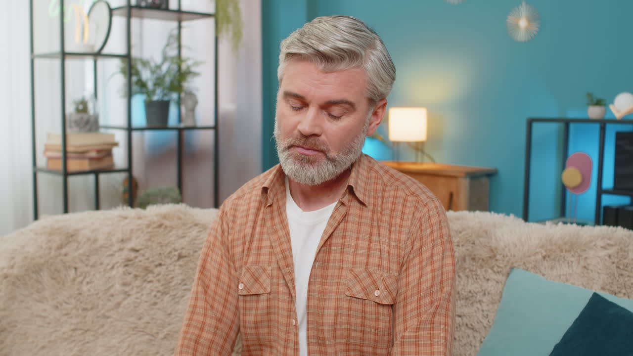 Mature man sitting on sofa holding smartphone expressing stress after receiving bad news at home