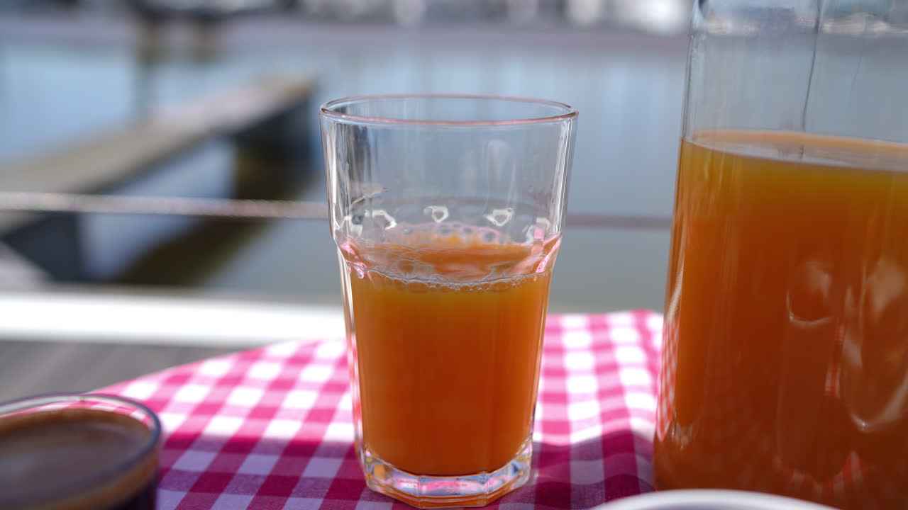 Pouring orange juice into glass on checkered tablecloth near lake