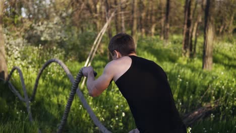 el deportista está entrenando con cuerdas onduladas. trabajo duro. camiseta negra. parque verde con árboles. sol. vista de atrás