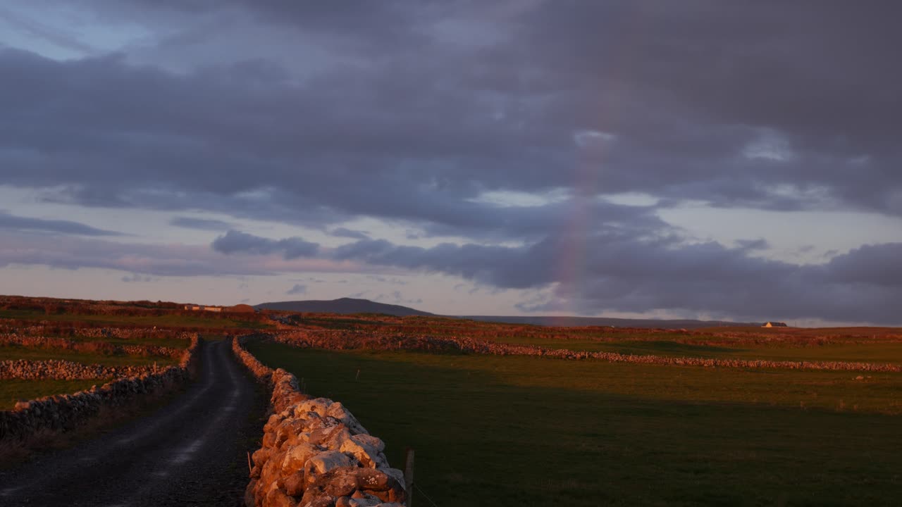 A winding country road lined with stone walls stretches through the Irish countryside under a dramatic sky, with soft evening light and a faint rainbow adding charm to the rural landscape