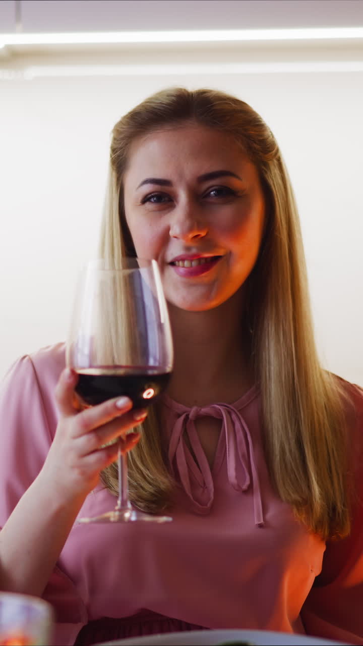 Seductive woman in pink dress cheers with wineglass and drinks vintage red wine sitting at table during romantic dinner at home slow motion