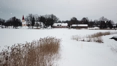 Village-church-and-wooden-homes-covered-in-white-snow-during-snowfall,-low-angle-drone-view