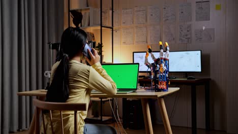 back view of teen asian girl talking on smartphone while designing a cyborg hand on a laptop with mock up green screen at home
