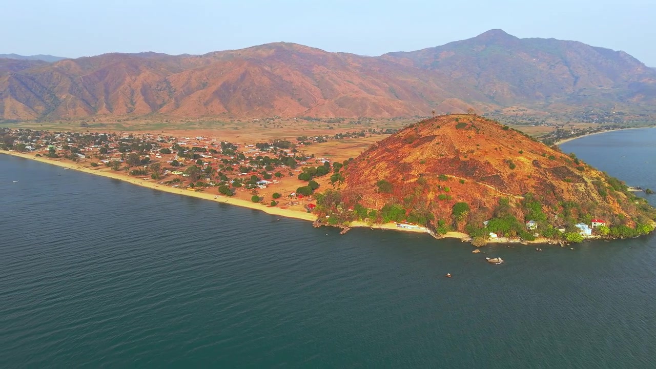 Un plan aérien en recul depuis la colline distinctive d'Usisya révèle le village lacustre, le lac Malawi bleu-vert, de petits bateaux et des montagnes au loin.