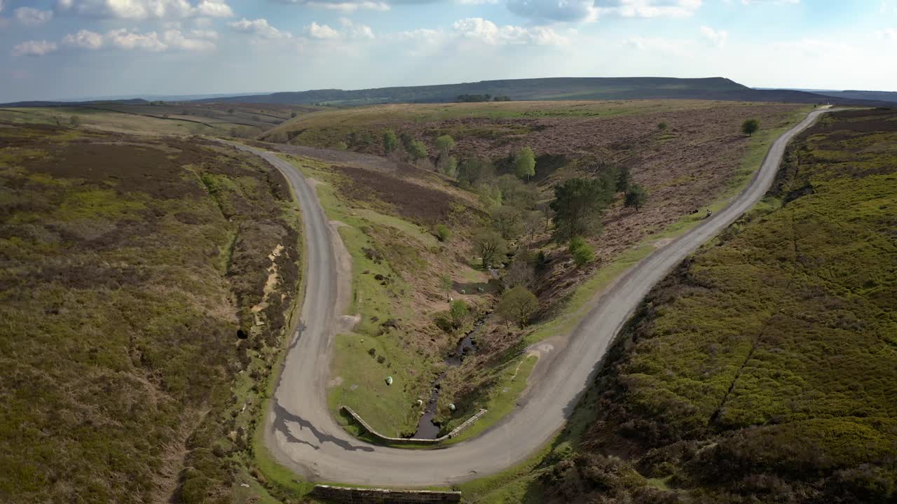 작은 다리 아래로 흐르는 황무지 개울을 보여주는 공중 비디오, 그리고 숲이 우거진 계곡을 통해 저지대로 향함