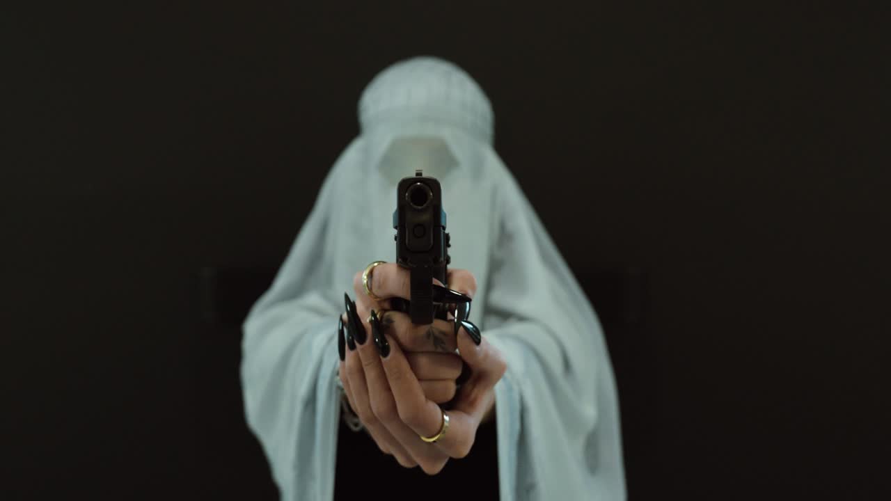 A woman dressed in traditional Muslim clothing poses against a dark background. She holds a pistol and points it at the camera