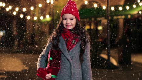 retrato de una linda chica feliz sosteniendo un regalo de navidad y sonriendo a la cámara en la calle mientras nieva en navidad