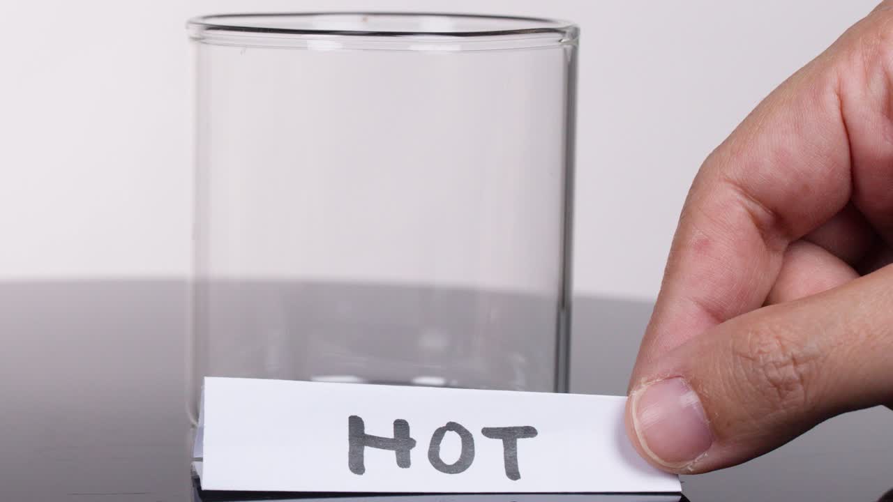 A hand places a 'HOT' label on a clear glass beaker in a studio setting