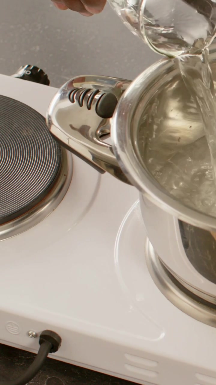 Vertical video: Pouring from glass cup hand filling steel pot on counter placing pot on hob to boil
