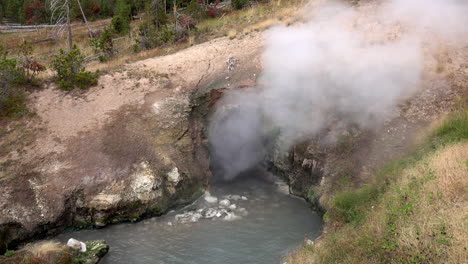 steam rises from dragon's mouth spring in yellowstone national park, and a wave of water is ejected from the opening