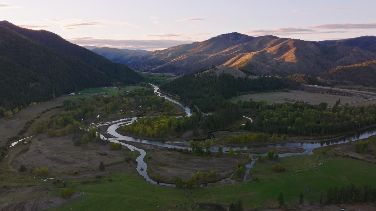 Serpentine river channel weaving through green rural valley bordered by forested hills and distant mountains, aerial drone, ascend