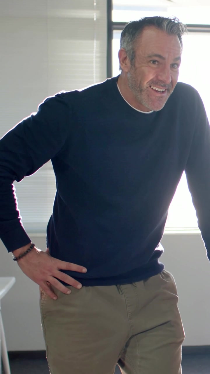 Vertical video: Leaning man smiling after offscreen prompt in office in navy top touching laptop