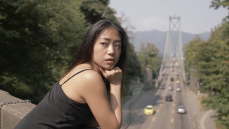 medium closeup of pretty asian woman in black looking behind, lions gate bridge in background, slowmo