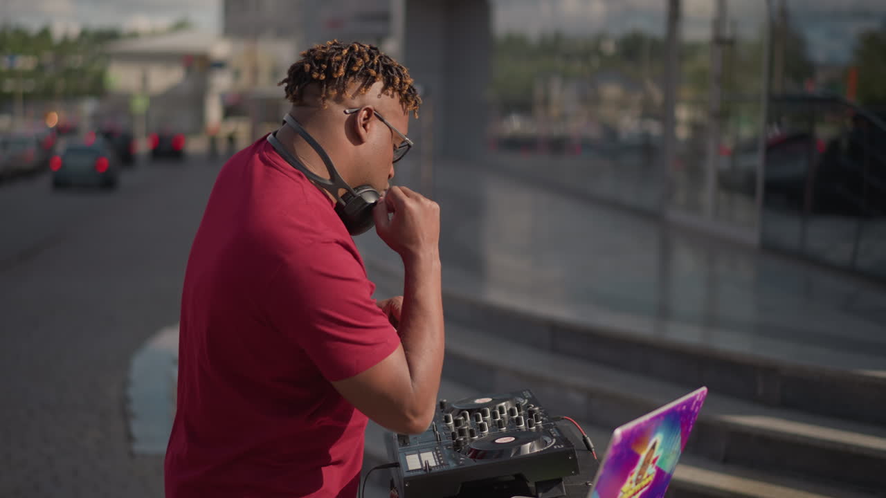 Deejay nero che mixa all'angolo della strada, cuffie per laptop, performance diurna energica, maglietta rossa, controller portatile, skin colorata per laptop, auto che passano, reazioni dei pedoni, mette in coda i brani, pugno alzato