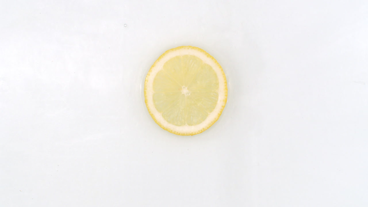 On a white background a splash of water falls on a slice of lemon in slow motion.