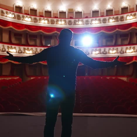 man in front of empty theatre hall with red velvet chairs. actor is standing on stage and talking with gestures before the performance