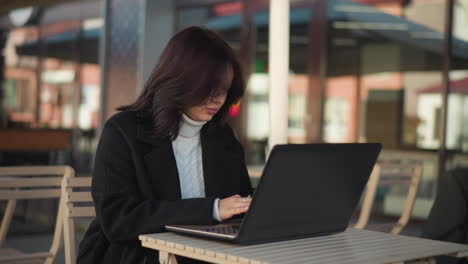 tech-savvy professional focused on laptop screen while seated outdoors in urban environment, scene features natural light, modern furniture, blurred background of building