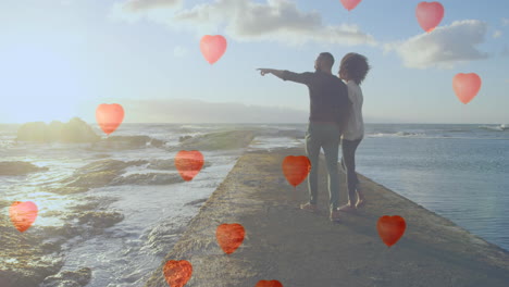 multiple heart balloons floating against couple pointing towards sea shore