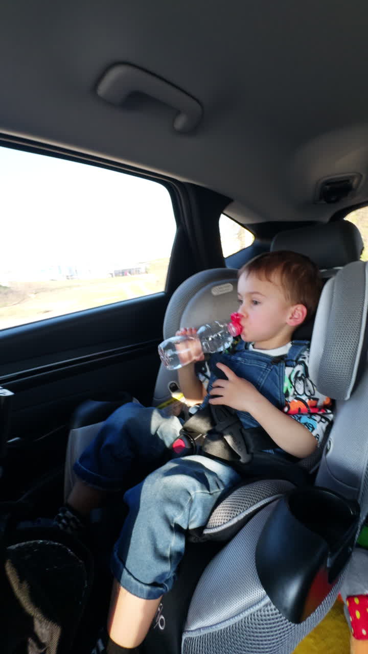 Calm toddler boy sitting comfortably in baby seat chair. Child drinking water from bottle during his travel. Vertical video.