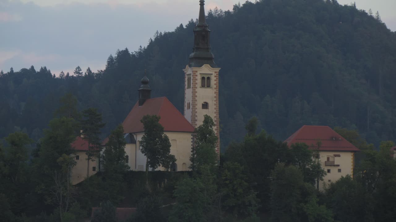 Inquadratura statica della chiesa sull'isola del Lago di Bled al crepuscolo a Bled, Slovenia. Le acque calme e le montagne boscose creano un'atmosfera di viaggio serena.