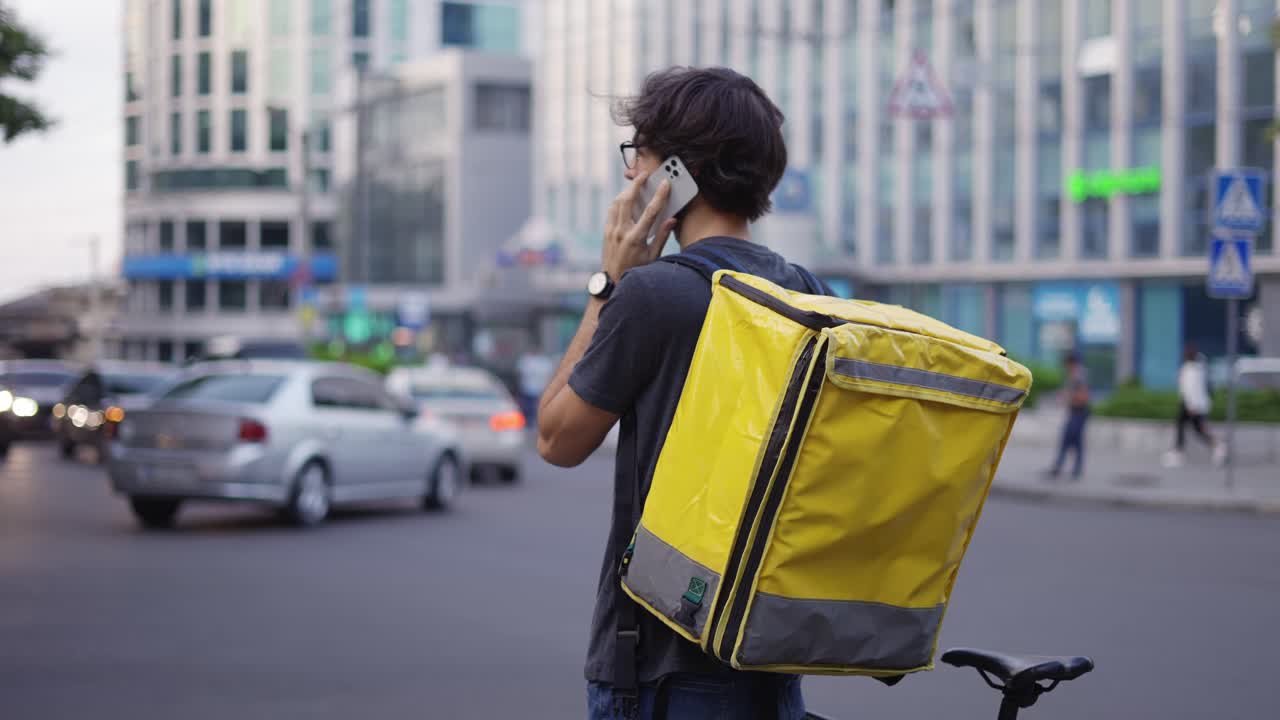 Delivery man with backpack calling to a client on the city street, rear view