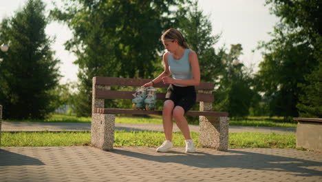 una joven blanca colocando sus patines de color cian en un banco de madera del parque antes de sentarse, lleva una pulsera negra y zapatillas blancas, con el fondo con árboles verdes vibrantes