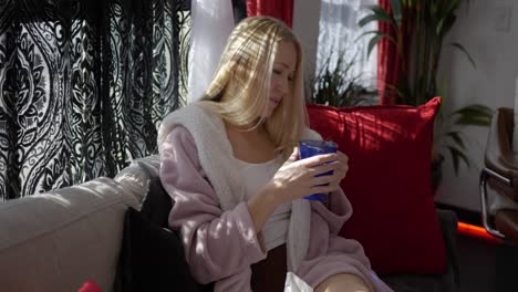 Beautiful-blonde-white-caucasian-girl-women-sitting-on-couch-in-front-of-windows-in-pink-bath-robe-resting-relaxing-at-home-with-hot-cup-of-tea-water-in-blue-glass-mug-blowing-on-steam-to-cool-down