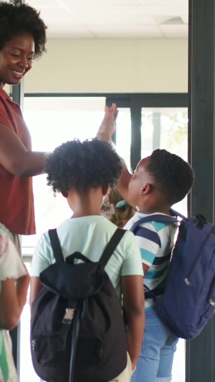 Vertical video: Greeting teacher rust top high-fiving students arriving with backpacks into room