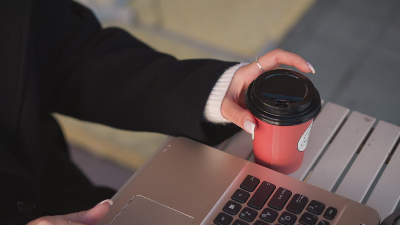 나무 테이블 위의 노트북 키보드 위에 커피 컵을 들고 있는 여자의 손의 클로즈업, 은 조명과 함께 은 손과 캐주얼 야외 작업 공간 설정을 강조합니다.