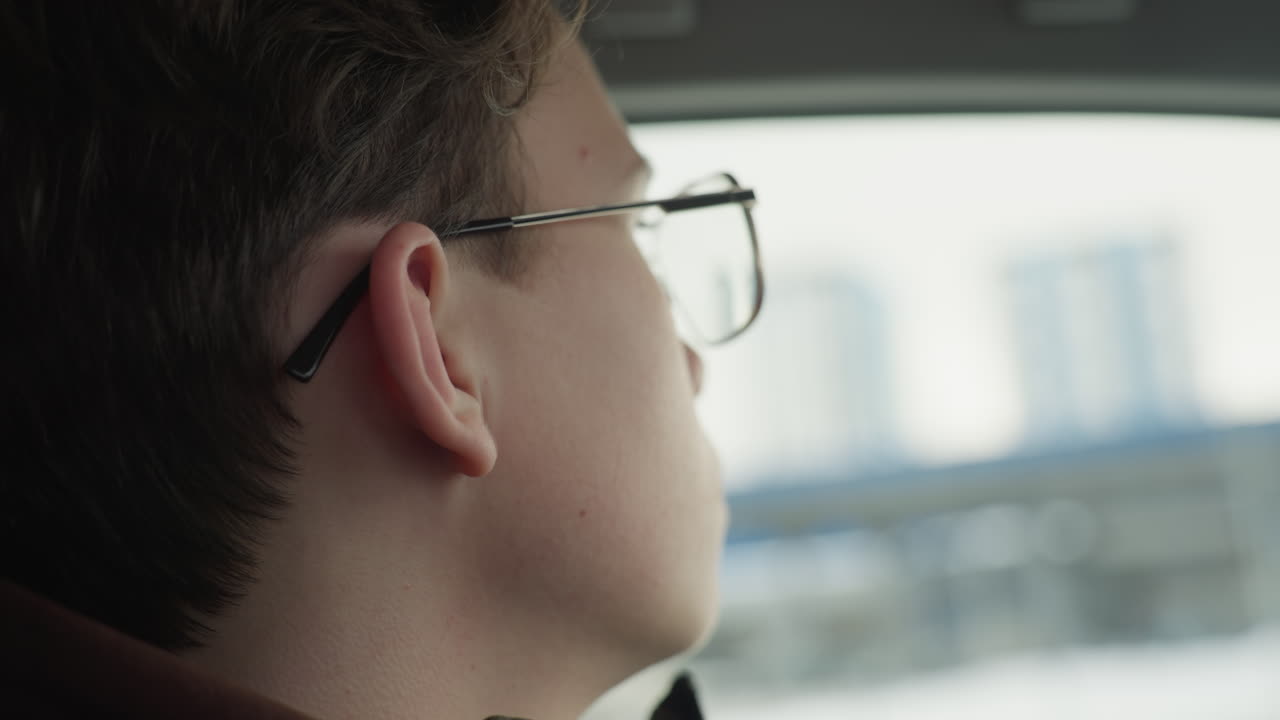 Close up of young man adjusting glasses with hand on steering wheel inside car cabin while blur snowy background visible through window capturing reflective gaze