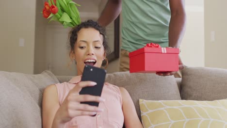 mujer biracial sorprendida sentada en el sofá con un teléfono inteligente recibiendo un regalo de un compañero masculino