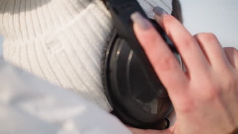 close up of black over ear headphone pod being adjusted by hand wearing white knit winter cap over brown hair, sunlight glinting on metal band under clear blue sky in cold urban setting