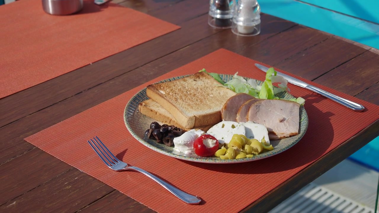 Frisk morgenmadsanretning med toast, skinke, feta og oliven på et solrigt udendørs bord