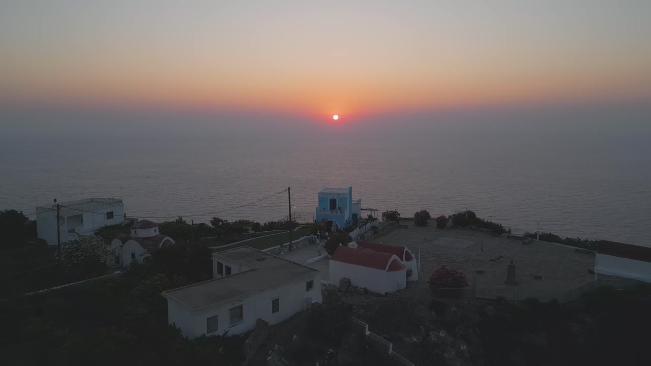 Drone reveal of Mesochori village square in Karpathos, a unique spot to enjoy a breathtaking Aegean Sea sunset as the horizon glows with warm evening colors