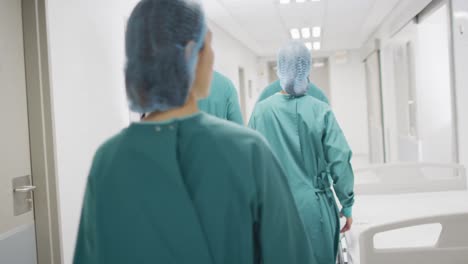 Rear-view-of-group-of-diverse-surgeons-in-surgical-caps-and-gowns-walking-in-hospital-corridor