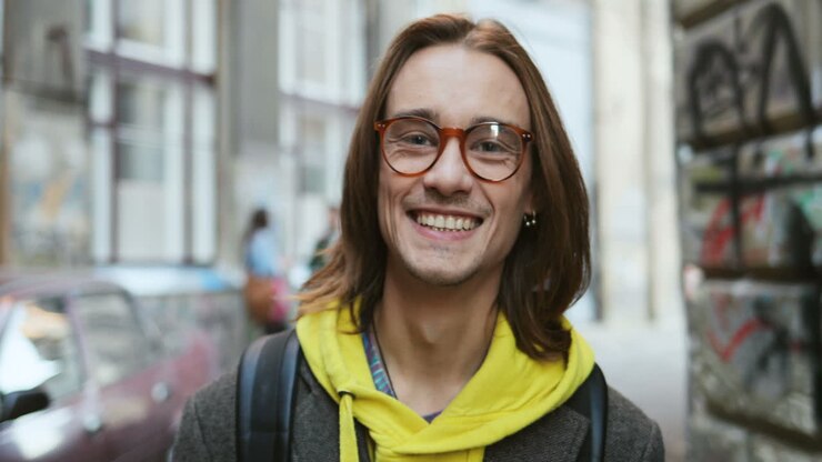Primo piano di un giovane uomo caucasico con i capelli lunghi e gli occhiali che sorride alla macchina fotografica per strada
