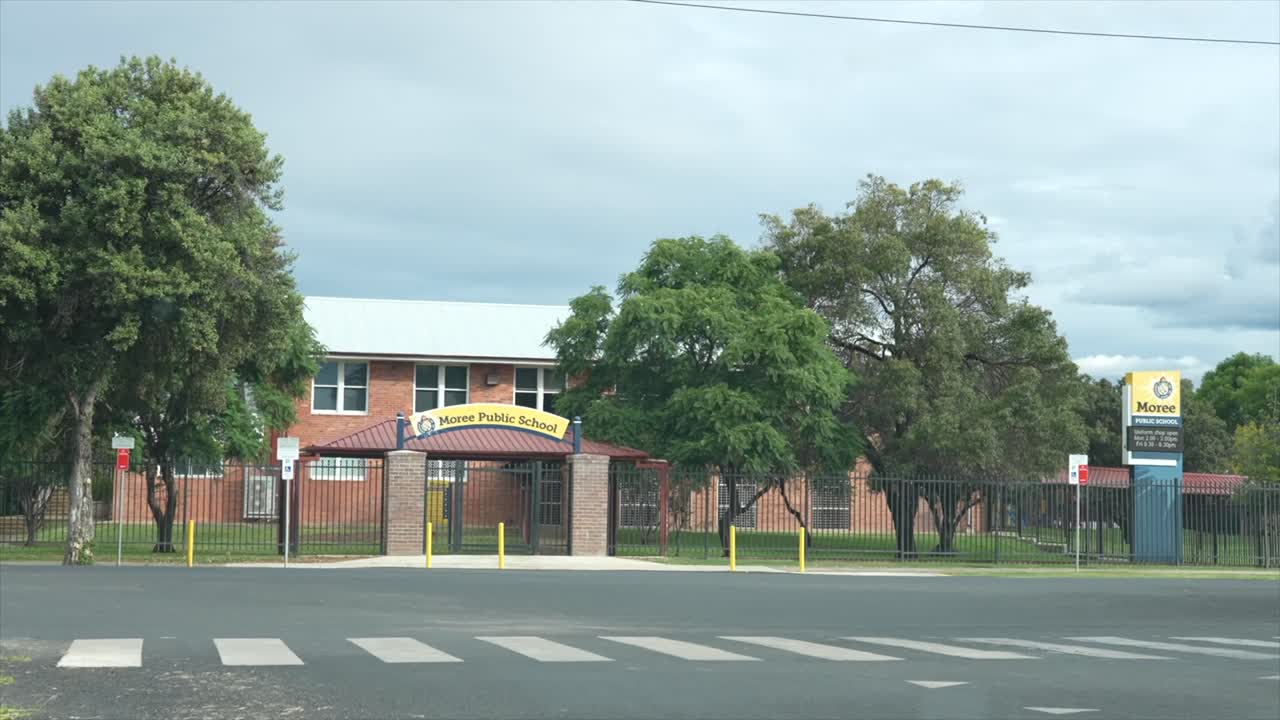 A quiet street view of Moree Public School in regional New South Wales, showing the front entrance, brick buildings, mature trees and a pedestrian crossing under overcast skies