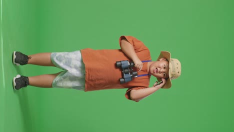 niño turista asiático con un sombrero y binoculares hablando en teléfono inteligente mientras está de pie en el fondo de la pantalla verde. niño investigador examina algo, concepto de aventura de turismo de viaje, cuerpo completo