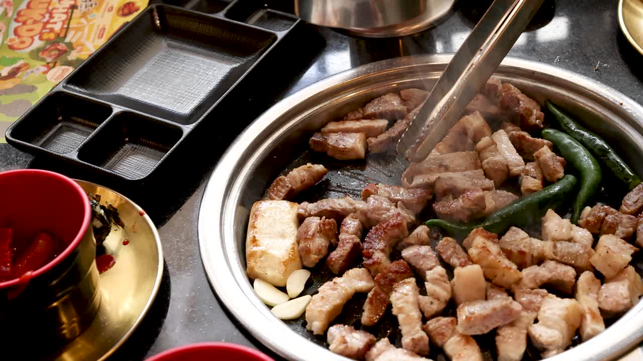 Tongs stir pork belly, garlic, and peppers on tabletop grill in bright, modern restaurant setting