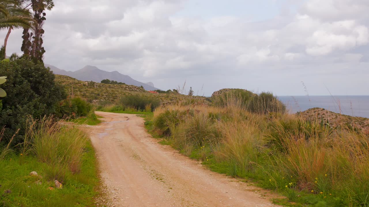 Picturesque view of unpaved road winding through landscape of dry, golden grasses and bushes under cloudy sky in Sicily, Italy