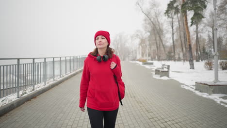 woman with headphones around neck walking along interlocked pathway in red hoodie with bag over shoulder, surrounded by snow-dusted park, iron railing, benches, and foggy winter atmosphere