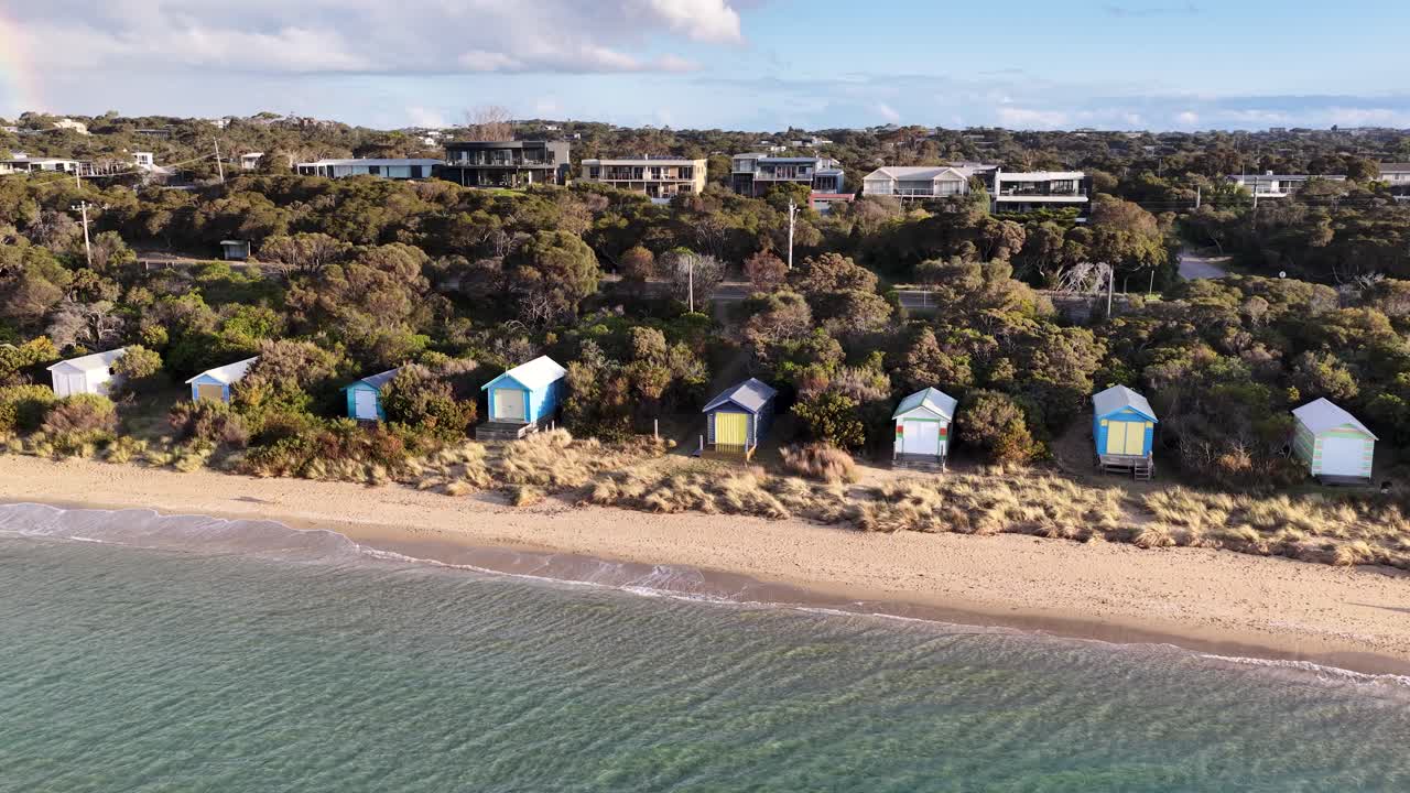 Drone pans above sandy beach, vibrant bathing boxes, and coastal vegetation in soft daylight