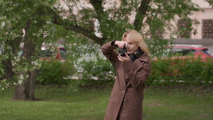 Lady stands outdoors holding digital camera framed from waist up. Woman wears long brown coat and has blonde hair. background features green trees with white flowers grassy ground distant