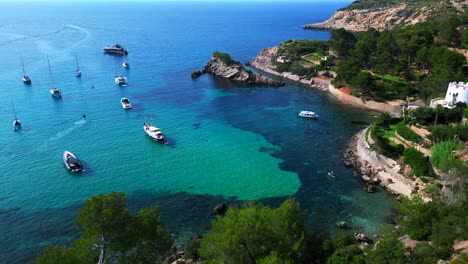 sailboats and yachts moored in turquoise waters of a bay on ibiza, spain, surrounded by lush greenery. majestic aerial view flight fly reverse drone