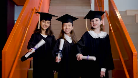 Three-Happy-Preschool-Female-Students-In-Cap-And-Gown-Showing-Their-Diplomas-And-Looking-At-The-Camera-At-The-Graduation-Ceremony