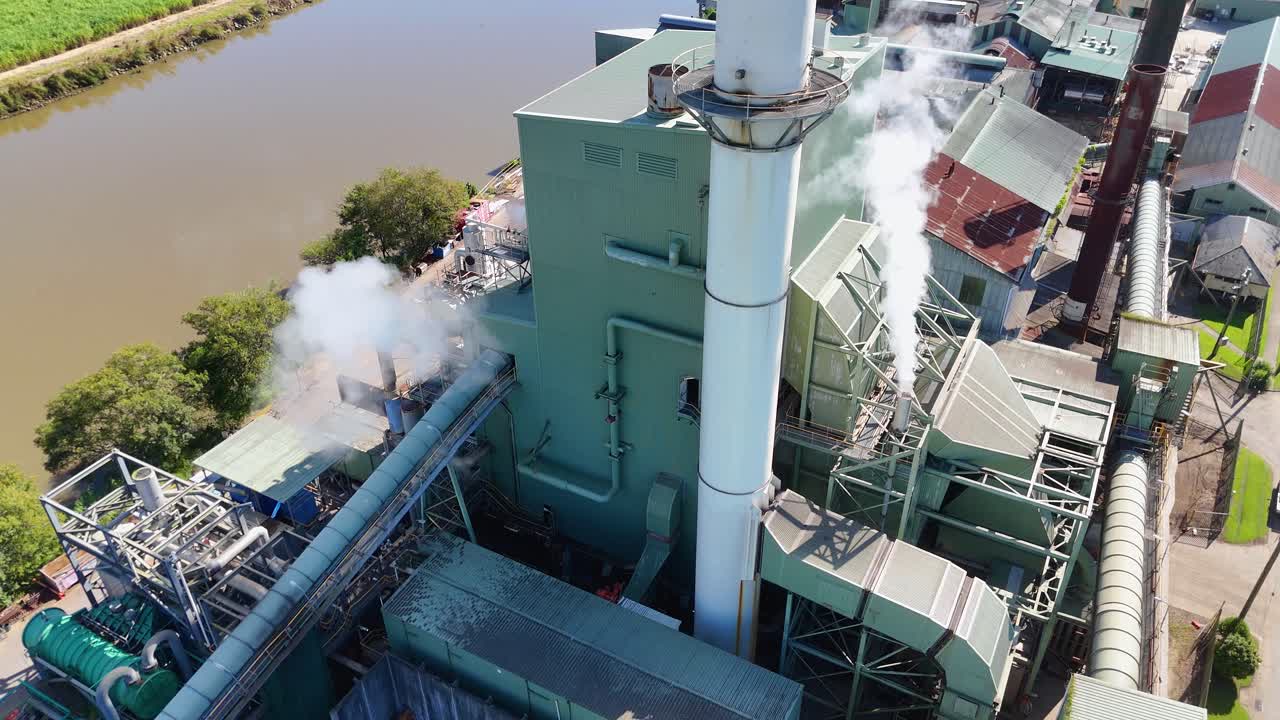 Drone footage captures a factory with smoke stacks near a river, highlighting industrial activity and environmental impact